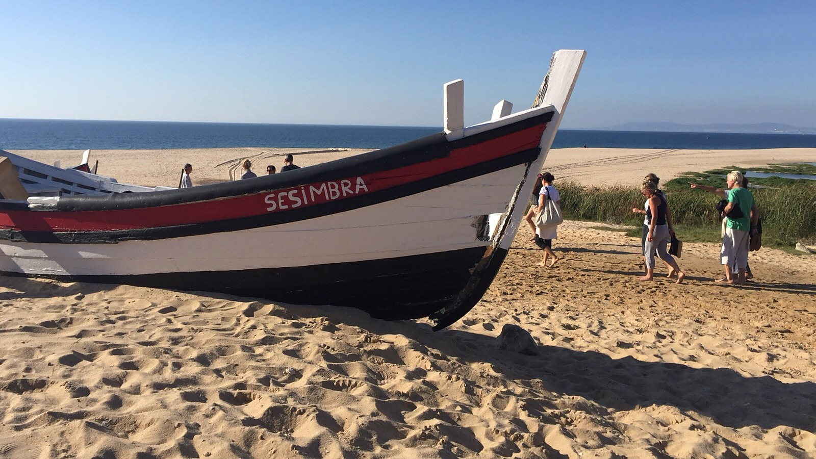 Une barque de pêche traditionnelle peinte en blanc, rouge et noir, tirée sur une plage de sable, avec la mer en arrière-plan et quelques personnes qui marchent