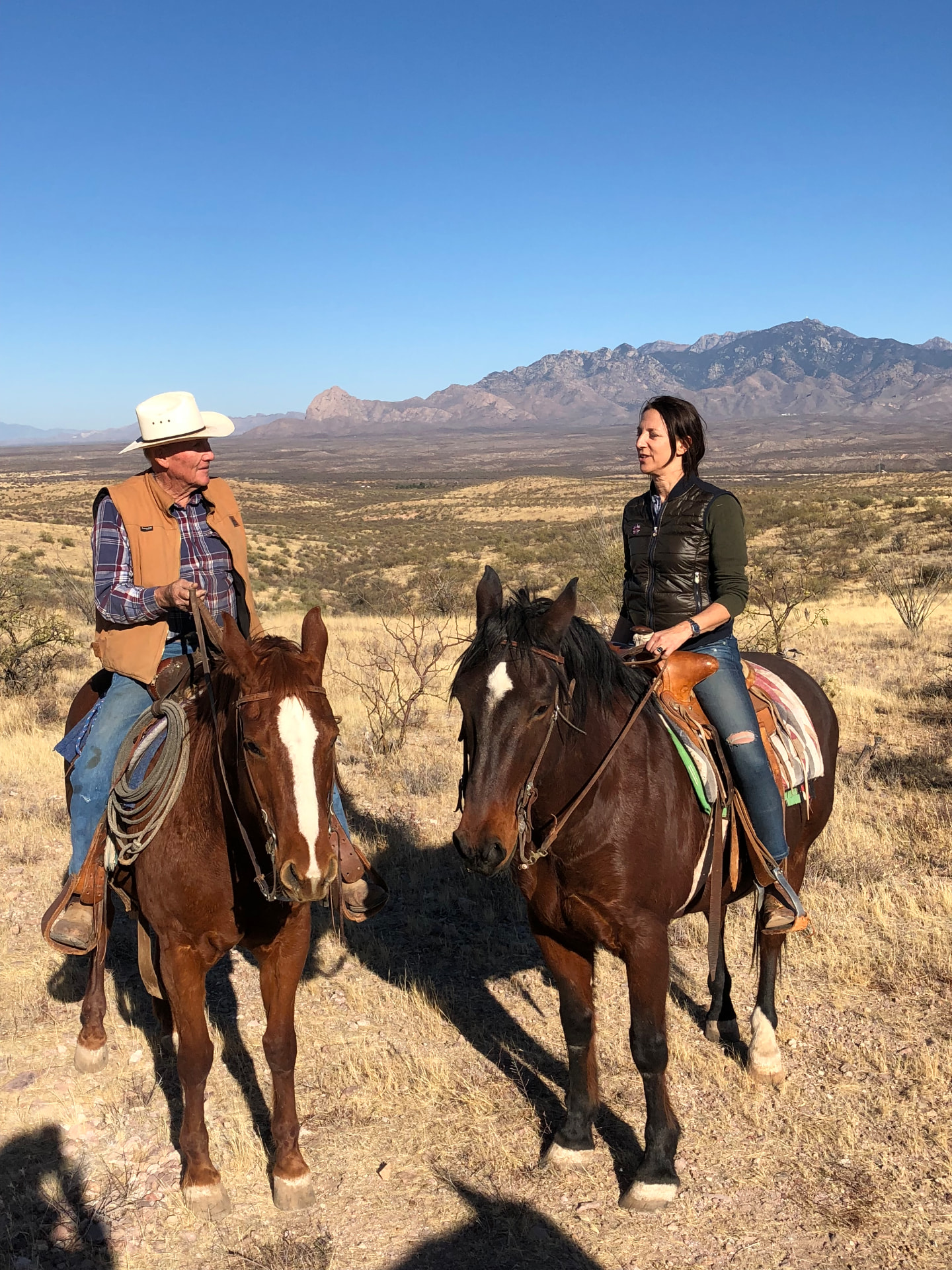 L'autrice à cheval en conversation avec un cow-boy dans un paysage désertique, montagnes en arrière-plan — Nogales, Arizona